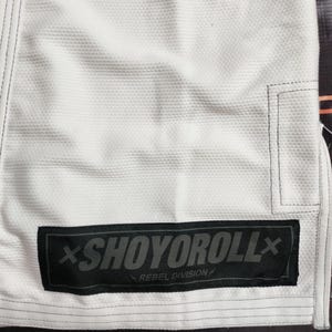 Op de afbeelding: Close-up van een wit vechtsportuniform met zwarte stiksels en een zwart rechthoekig embleem. Het embleem toont de grijze tekst "SHOYOROLL" en de woorden "REBEL DIVISION" eronder.