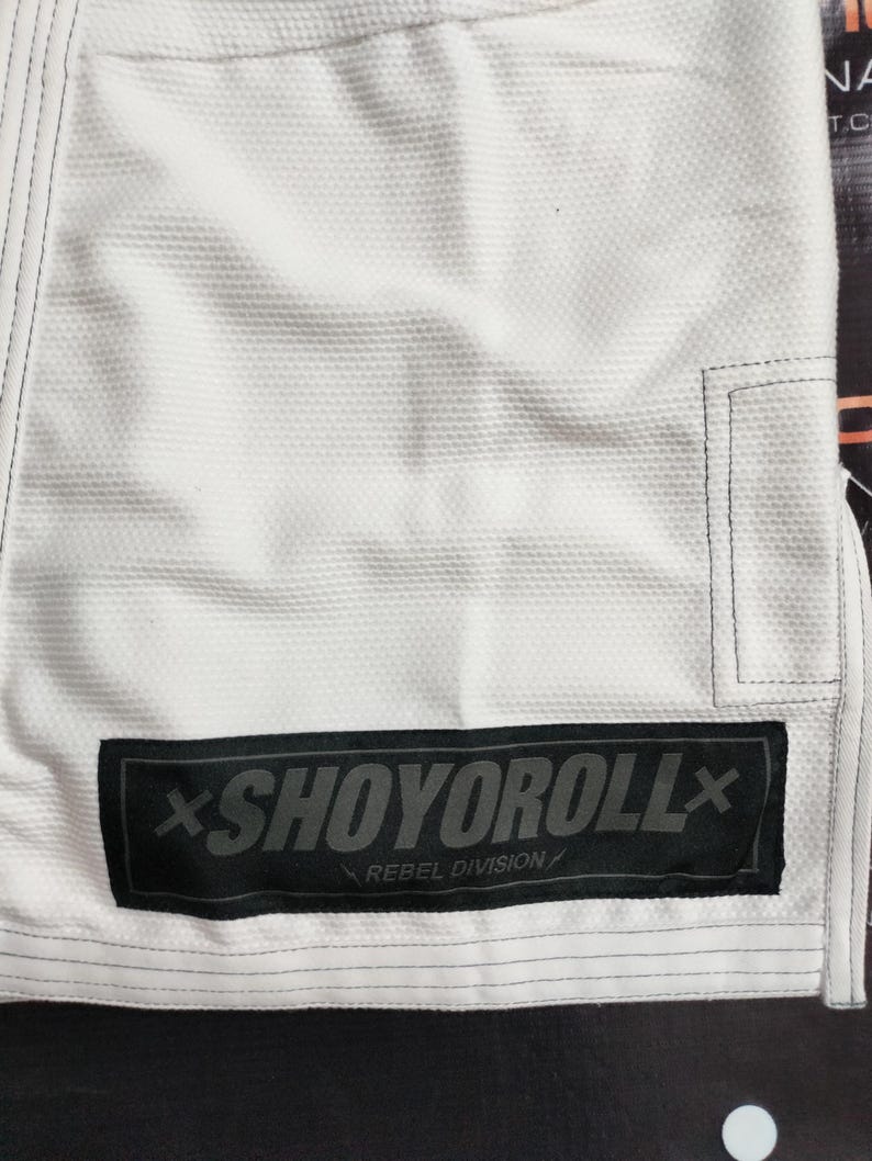 Op de afbeelding: Close-up van een wit vechtsportuniform met zwarte stiksels en een zwart rechthoekig embleem. Het embleem toont de grijze tekst "SHOYOROLL" en de woorden "REBEL DIVISION" eronder.