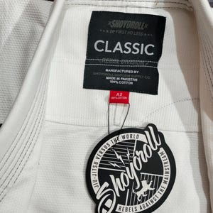 Op de afbeelding: Witte Jiu-Jitsu uniformjas met een geweven textuur. Het label luidt "CLASSIC" en "SHOYOROLL". Een zwart-wit label met de tekst "JIU-JITSU VERSUS THE WORLD" en "REBELS AGAINST THE MAINSTREAM" is bevestigd.