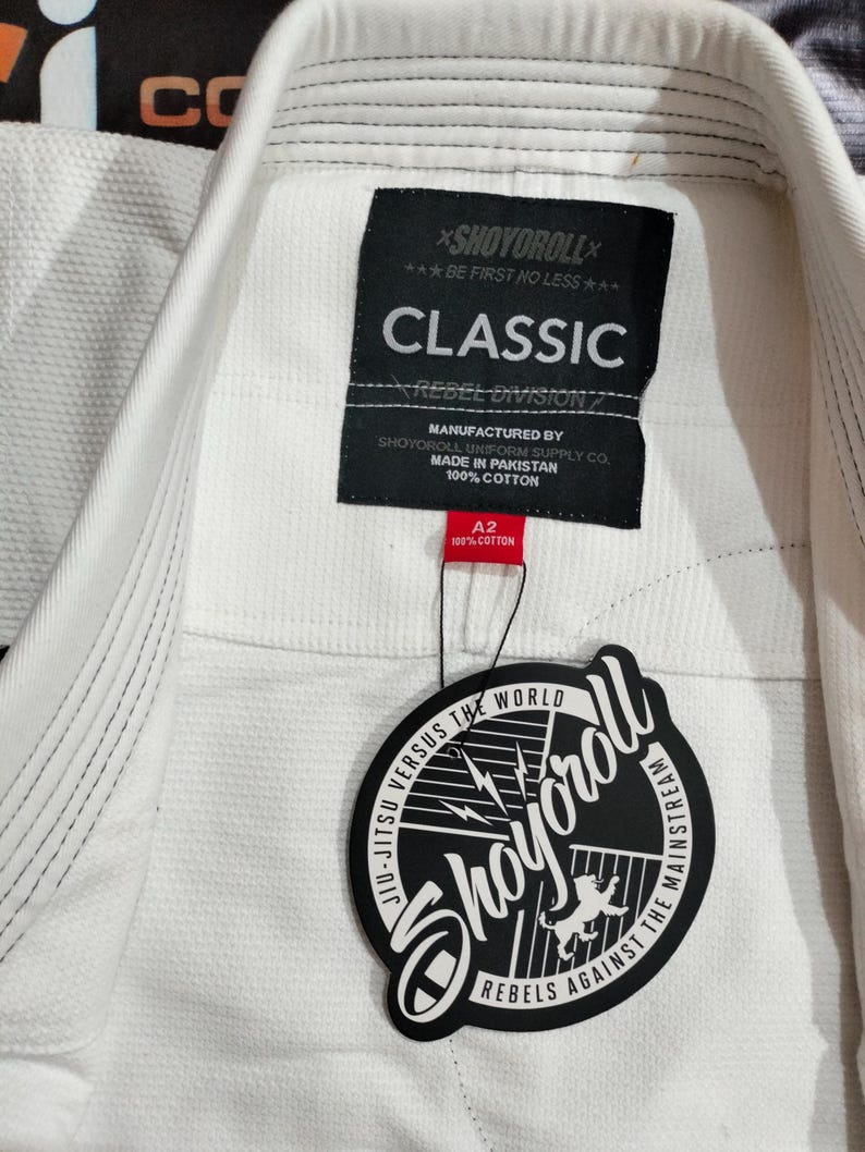 Op de afbeelding: Witte Jiu-Jitsu uniformjas met een geweven textuur. Het label luidt "CLASSIC" en "SHOYOROLL". Een zwart-wit label met de tekst "JIU-JITSU VERSUS THE WORLD" en "REBELS AGAINST THE MAINSTREAM" is bevestigd.