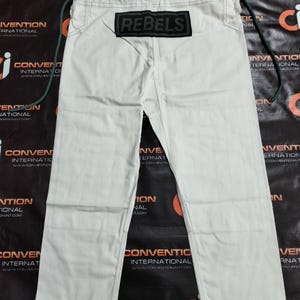 Op de afbeelding: Gebroken witte vechtsportbroek met het woord "REBELS" op een zwart label. De broek heeft een trekkoord en zwarte stikseldetails. De broek ligt plat.