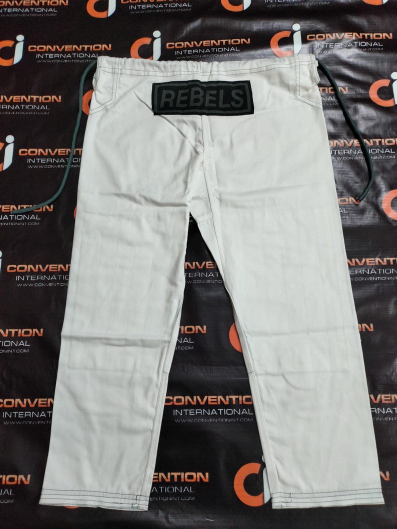 Op de afbeelding: Gebroken witte vechtsportbroek met het woord "REBELS" op een zwart label. De broek heeft een trekkoord en zwarte stikseldetails. De broek ligt plat.