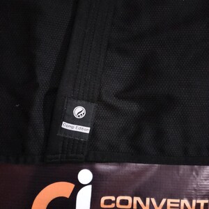 May include: Close-up of a black martial arts uniform with a textured weave. A small label reads "Comp Edition" and features a circular design. The bottom of the image shows a banner with the words "CONVENTION INTERNATIONAL".