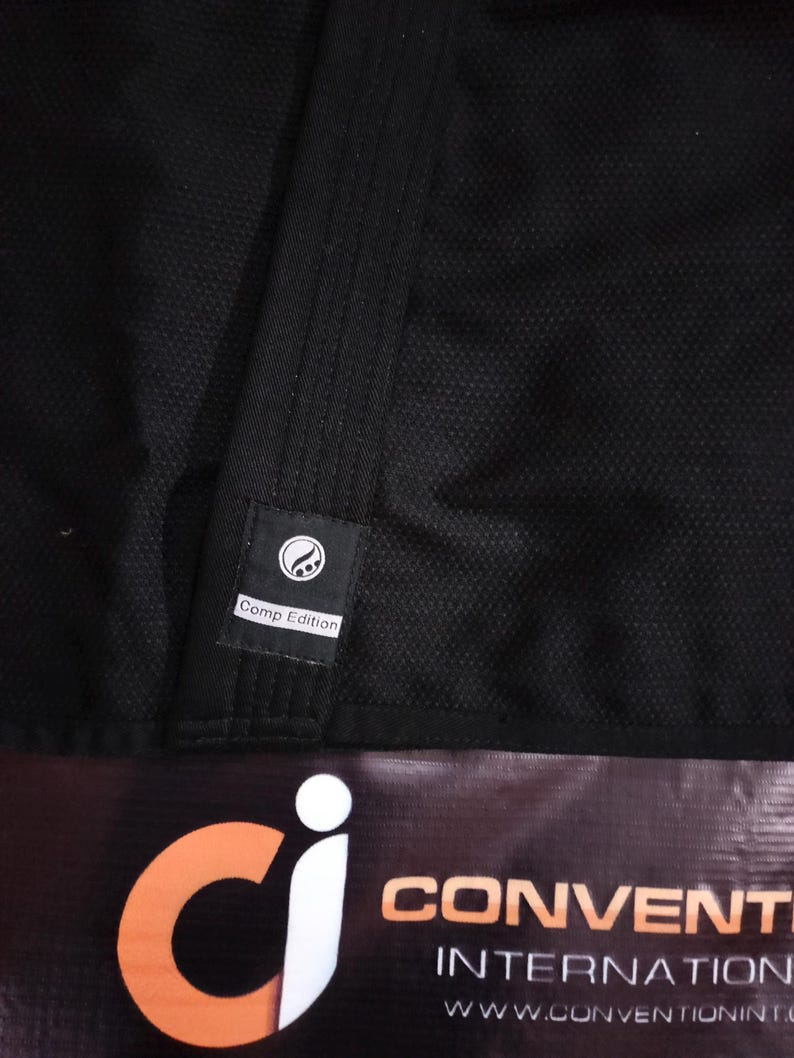May include: Close-up of a black martial arts uniform with a textured weave. A small label reads "Comp Edition" and features a circular design. The bottom of the image shows a banner with the words "CONVENTION INTERNATIONAL".