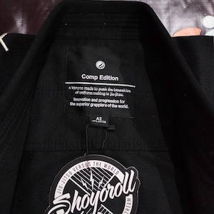 May include: Black jiu-jitsu gi with a tag that reads "Shoyoroll" and "Comp Edition." The gi has a collar and is made of cotton. The tag also states "Jiu-Jitsu Versus The World."