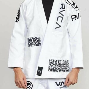 Puede incluir: Uniforme blanco de artes marciales con texto gráfico y logotipos negros. La chaqueta presenta el nombre de la marca "RVCA" en letras grandes y varios diseños abstractos. El uniforme incluye chaqueta y pantalón, adecuado para entrenamiento de artes marciales.