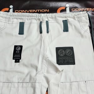 Op de afbeelding: Witte Jiu-Jitsu broek met donkergrijze accenten. Voorzien van een Shoyoroll Jiu Jitsu patch en een kleinere patch met Japanse tekst. De broek heeft versterkte stiksels en riemlussen.