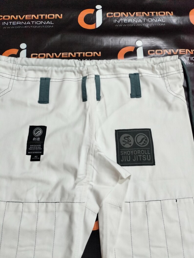 Op de afbeelding: Witte Jiu-Jitsu broek met donkergrijze accenten. Voorzien van een Shoyoroll Jiu Jitsu patch en een kleinere patch met Japanse tekst. De broek heeft versterkte stiksels en riemlussen.
