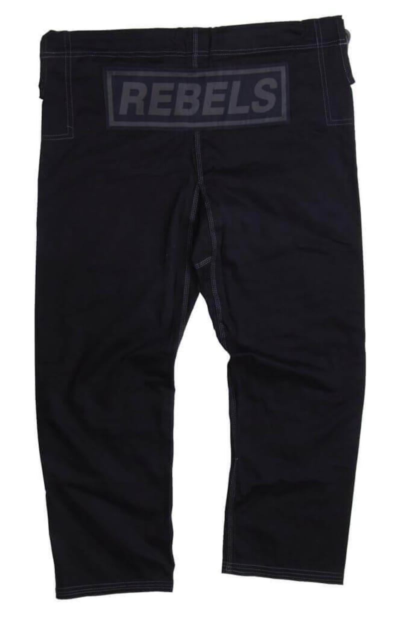 Op de afbeelding: Zwarte vechtsportbroek met het woord "REBELS" in donkergrijze letters op de rug. De broek heeft witte stiksels en een relaxte pasvorm. De broek is gemaakt van een duurzaam materiaal.