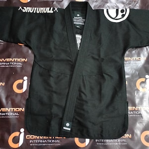 May include: A black martial arts gi with white lettering and a circular logo. The gi features a traditional collar, reinforced seams, and a tailored fit. The gi is designed for martial arts training and competition.