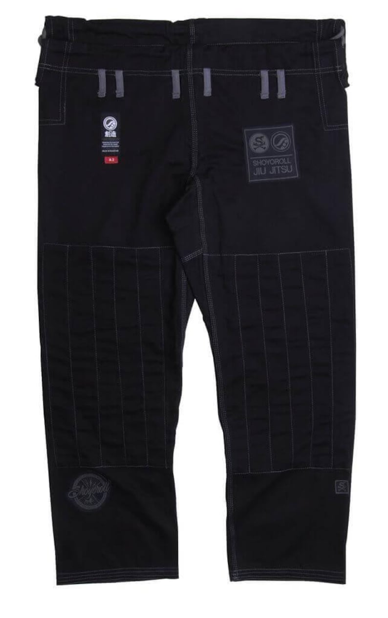Op de afbeelding: Zwarte Jiu-Jitsu broek met versterkte stiksels en patches. De broek heeft het "Shoyoroll Jiu Jitsu" logo en andere merkemblemen. Het ontwerp bevat grijze accenten en een getailleerde pasvorm, geschikt voor vechtsporttraining.