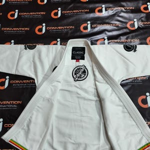Op de afbeelding: Wit martial arts uniform, waarschijnlijk een gi, plat neergelegd. De jas is open en toont de binnenvoering. Een label leest "CLASSIC" en een cirkelvormig logo is zichtbaar. De rand heeft rode, gele en groene strepen. De achtergrond is zwart met herhaalde logo's.