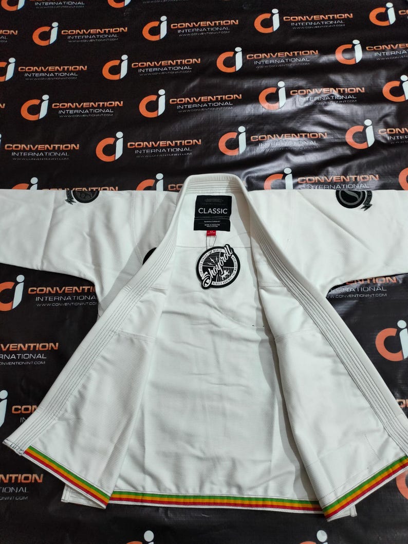Op de afbeelding: Wit martial arts uniform, waarschijnlijk een gi, plat neergelegd. De jas is open en toont de binnenvoering. Een label leest "CLASSIC" en een cirkelvormig logo is zichtbaar. De rand heeft rode, gele en groene strepen. De achtergrond is zwart met herhaalde logo's.