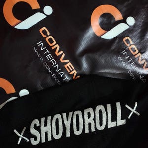 May include: Black fabric with the white embroidered text "SHOYOROLL X". The background features a black material with orange and white logos and the text "CONVENTION INTERNATIONAL".