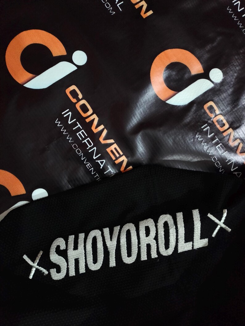 May include: Black fabric with the white embroidered text "SHOYOROLL X". The background features a black material with orange and white logos and the text "CONVENTION INTERNATIONAL".