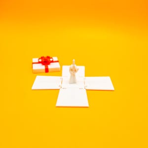 May include: A white gift box with a red ribbon is open and a white hand with the middle finger extended is inside the box.