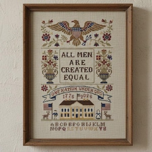 May include: Framed cross-stitch artwork featuring an American eagle, floral motifs, and the text "ALL MEN ARE CREATED EQUAL" and "ONE NATION UNDER GOD 1776 2026." The artwork includes a building, flags, and an alphabet.
