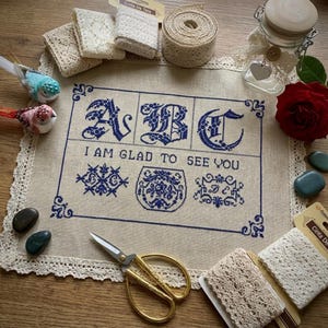 May include: A cross-stitched sampler with the letters ABC and the phrase "I AM GLAD TO SEE YOU" in blue thread. The sampler is surrounded by cream-colored lace trim. Additional items include lace ribbon, decorative birds, scissors, and a red rose.