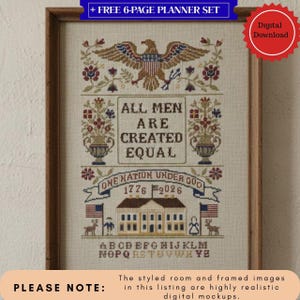 May include: Framed cross-stitch sampler with a patriotic theme. The design includes an eagle, floral elements, and the text "ALL MEN ARE CREATED EQUAL." Additional text includes "ONE NATION UNDER GOD 1776 2026." The sampler is in a wooden frame.
