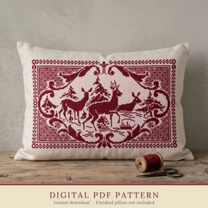 May include: A rectangular throw pillow featuring a deep red cross-stitch pattern on a cream fabric. The design depicts three deer standing among pine trees, enclosed within an ornate, decorative border. A spool of red thread and scissors lie nearby.