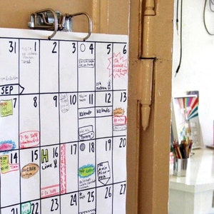 May include: A white calendar with black grid lines and handwritten appointments for September. The calendar is hanging on a door with a metal clip.