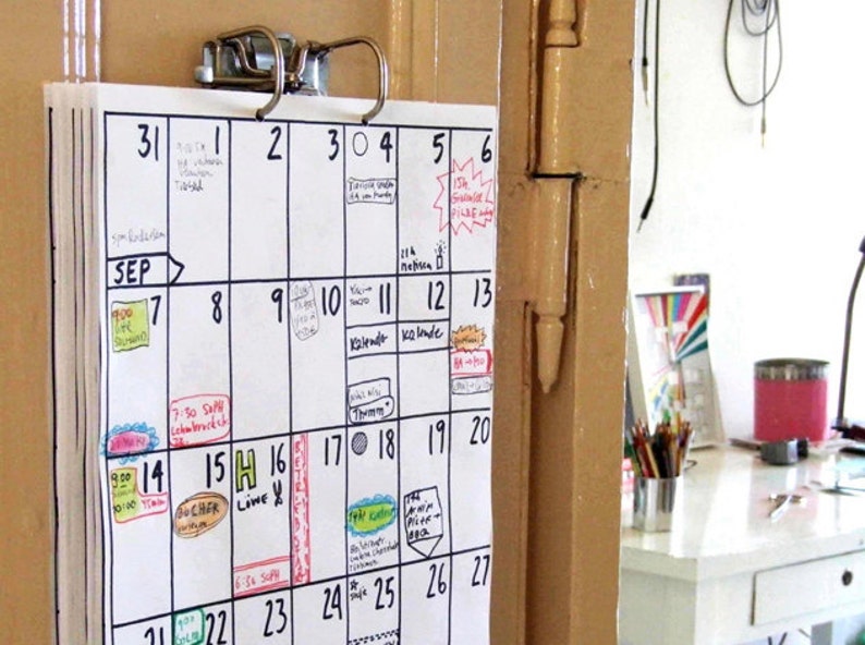 May include: A white calendar with black grid lines and handwritten appointments for September. The calendar is hanging on a door with a metal clip.