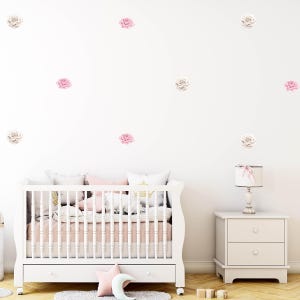 Könnte beinhalten: Ein weißes Babybett mit rosa und weißer Bettwäsche, ein weißer Nachttisch und eine Lampe mit einer rosa Schleife. Die Wand ist mit rosa und weißen Rosen-Wandtattoos dekoriert. Der Raum hat eine weiche, feminine Ästhetik.