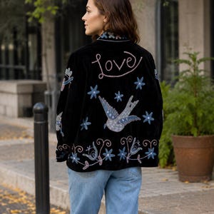 May include: A black jacket featuring embroidered designs on the back, including the word love in cursive script, a central bird, and various blue and pink floral motifs. The jacket has a collar and is worn with light blue denim jeans.
