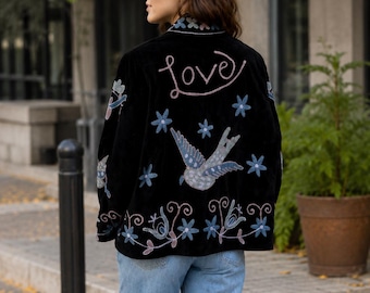 May include: A black jacket featuring embroidered designs on the back, including the word love in cursive script, a central bird, and various blue and pink floral motifs. The jacket has a collar and is worn with light blue denim jeans.