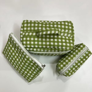 May include: Three green and white gingham patterned cosmetic bags. The largest bag has a handle and a quilted design. The bags have white zippers and are arranged on a white surface. The bags are a light green colour.