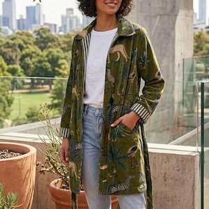 May include: An olive green long coat with a jungle animal print, including zebras and leopards. The coat has a striped lining and belt, worn with a white t-shirt and blue jeans. The person is wearing white trainers.