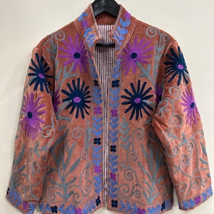 May include: A coral-colored jacket with intricate floral embroidery in shades of purple, blue, and green. The jacket features a velvet texture and a striped lining. The design includes large stylized flowers and scrolling vines.