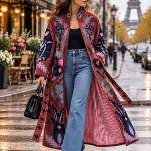 May include: A long, rust-colored coat featuring intricate floral embroidery in shades of pink, blue, and dark navy. The coat is worn over a black top and blue denim jeans, paired with a black handbag and black boots.