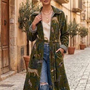 May include: A long, olive green coat with a jungle animal print, worn over blue jeans. The coat features a belt, pockets, and striped trim. The model is wearing brown boots.