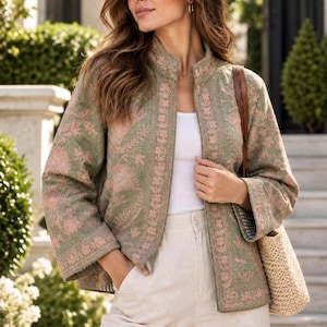 Op de afbeelding: Een saliegroene jas met een mandarijnkraag en lange mouwen, versierd met ingewikkeld roze bloemenborduursel. De jas wordt gedragen over een effen wit topje en een lichtbeige broek, gecombineerd met een geweven schoudertas van stro.
