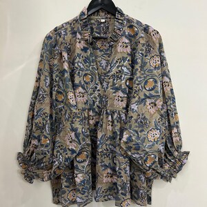 May include: A long-sleeved blouse in a muted olive green base colour featuring a repeating floral pattern in shades of navy blue, mustard yellow, and soft pink. The garment has a ruffled collar, a V-neckline, and gathered cuffs at the wrists.