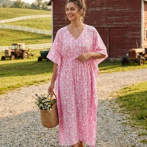May include: A loose-fitting kaftan dress featuring a vibrant pink and white abstract pattern, a V-neckline, and a drawstring waist. The garment has wide, short sleeves and falls to a midi length, paired with brown sandals and a woven straw basket.