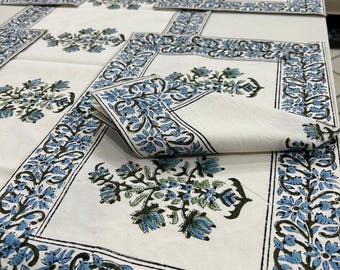Op de afbeelding: Een witte tafelkleed en bijpassende placemats en servetten met een blauw bloemmotief. De rand en de bloemstukken hebben blauwe en groene bloemen en bladeren. De tafelsetting is op een wit oppervlak geplaatst.