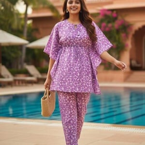 May include: A woman wearing a matching purple floral print top and pants set. The top has a drawstring waist and short sleeves. She is carrying a woven handbag and wearing white sandals. Sunglasses are on top of her head.