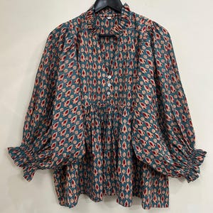 以下が含まれることがあります： 赤とクリーム色のティアドロップ柄が繰り返される、ティールカラーの長袖ブラウス。フリル付きのスタンドカラー、3つの白いボタンが付いたVネック、ギャザーの入った肩、そしてフリル付きの袖口で終わるボリュームのある袖が特徴です。