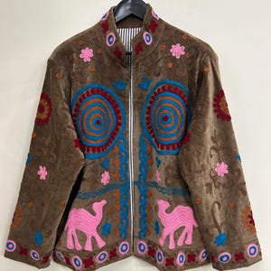 May include: A brown jacket featuring colorful embroidery, including pink camel motifs, blue spiral patterns, and various floral designs. The jacket has a high collar, a front zipper closure, and a black and white striped lining visible at the neck.