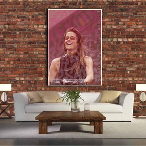 Può includere: Un poster incorniciato con una donna sorridente con occhiali da sole e cuffie. Il poster ha uno sfondo rosa e bianco con il testo "CHARLOTTE DE WITTE" in verticale a destra. È appeso a un muro di mattoni.