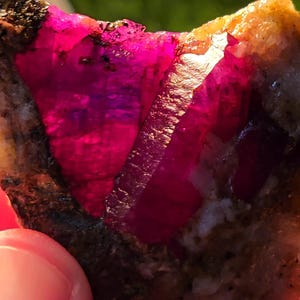 Pode incluir: Um close-up de um cristal de rubi rosa incrustado em uma rocha marrom áspera. O cristal é facetado e tem uma cor profunda e rica.