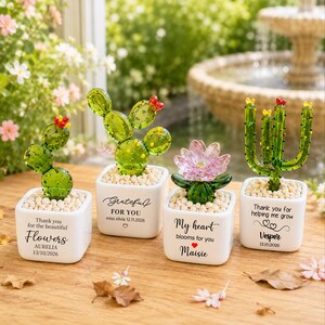 Pode incluir: Quatro figuras de plantas em vidro em vasos quadrados brancos com pequenas pedras. Os vasos apresentam o texto: "Thank you for the beautiful Flowers AURELIA 13/10/2026", "Grateful FOR YOU miss silvia 12.11.2026", "My heart blooms for you Maisie" e "Thank you for helping me grow Vesper 13.10.2026".