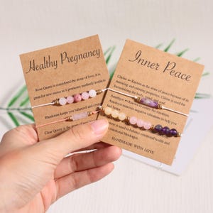 Puede incluir: Dos pulseras de piedras preciosas hechas a mano con tarjetas que dicen "Embarazo saludable" y "Paz interior". Las pulseras presentan cuentas rosas, moradas y transparentes. Las tarjetas brindan información sobre las piedras.