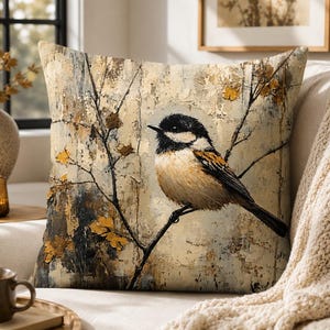 May include: A square throw pillow featuring a printed illustration of a small bird with a black head and tan chest perched on a thin, dark branch. The background has a textured, weathered appearance in shades of cream, beige, and dark brown.