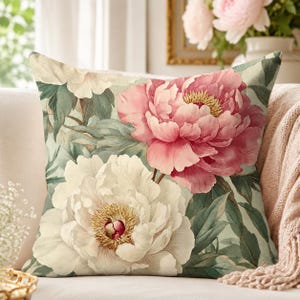 Könnte beinhalten: Ein quadratisches Zierkissen mit einem botanischen Druck aus großen, geschichteten rosa und weißen Pfingstrosen mit gelben Blütenmitten, umgeben von gedämpftem grünem Blattwerk auf einem hellen, neutralen Stoff.