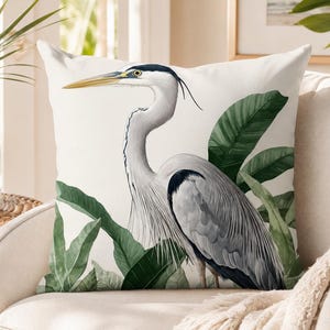 K&ouml;nnte beinhalten: Ein quadratisches Zierkissen mit einer detaillierten Illustration eines Graureihers, der zwischen gro&szlig;en gr&uuml;nen Tropenbl&auml;ttern steht. Der Vogel hat einen langen gelben Schnabel, einen wei&szlig;en Hals mit schwarzen Abzeichen und graue Federn.