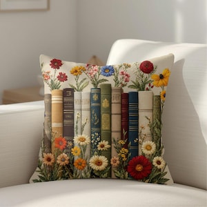 May include: A square throw pillow featuring a printed design of a row of vintage-style books standing upright. The books are surrounded by colorful illustrations of various wildflowers, including daisies, poppies, and sunflowers, in shades of red, yellow, and blue.