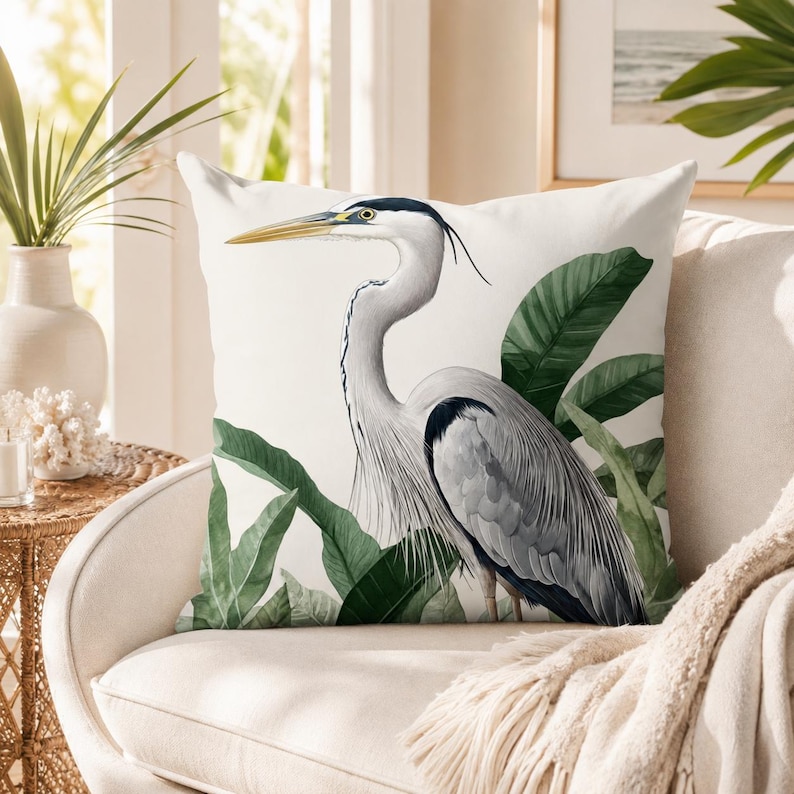 K&ouml;nnte beinhalten: Ein quadratisches Zierkissen mit einer detaillierten Illustration eines Graureihers, der zwischen gro&szlig;en gr&uuml;nen Tropenbl&auml;ttern steht. Der Vogel hat einen langen gelben Schnabel, einen wei&szlig;en Hals mit schwarzen Abzeichen und graue Federn.